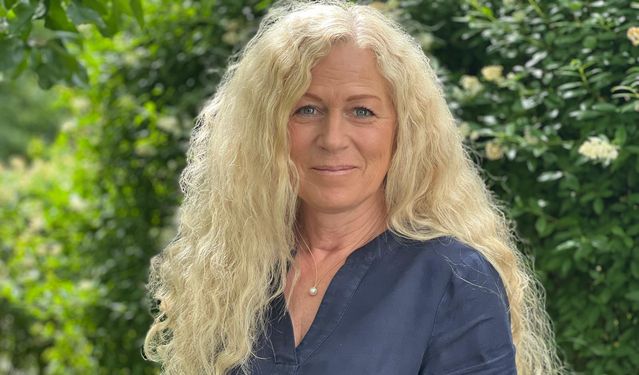 M.Sc. Psych. Katrin Bonato-Ehrenthal mit langen, lockigen, blonden Haaren in dunkelblauer Bluse, stehend vor einem blühenden grünen Hintergrund im Freien – mit freundlichem und professionellem Ausdruck.