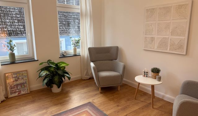 Modernes Wohnzimmer mit minimalistischer Einrichtung, bestehend aus einem grünen Sofa mit neutralen Zierkissen, einem runden Holzcouchtisch mit einer Keramikvase, einem beigefarbenen Sessel und Zimmerpflanzen. Natürliches Licht fällt durch große Fenster mit taupefarbenen Vorhängen und beleuchtet den beigefarbenen Teppich auf dem Holzboden.